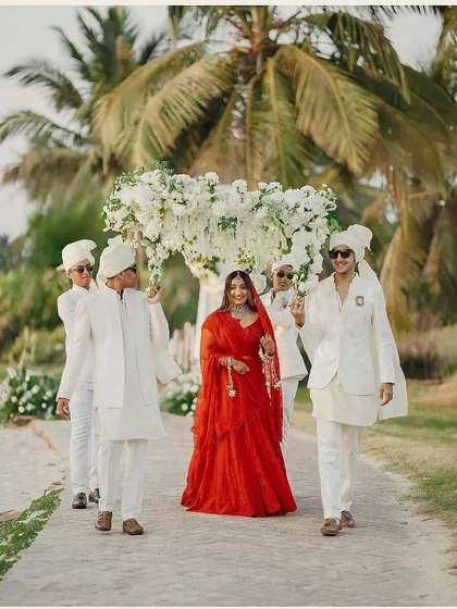 The bride's grand entrance with her groomsmen at a beautiful beach wedding in Goa. Her red lehenga stands out against the white decor, and her makeup is fresh and luminous for the daytime ceremony.