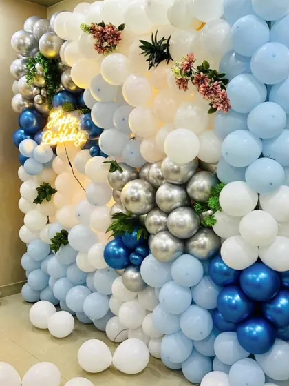 A stunning floor-to-ceiling balloon wall in shades of blue, white, and silver. A "Happy Birthday" neon sign is nestled within the balloons for a cool effect.