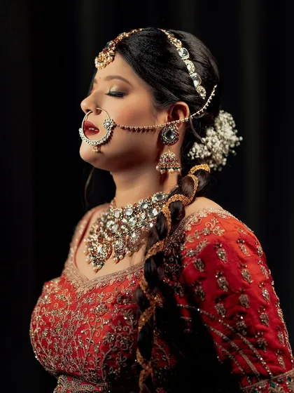 A serene and artistic bridal portrait. The bride's eyes are closed, drawing attention to her stunning makeup, traditional nath, and elaborate jewelry against a simple black backdrop.