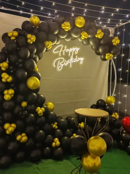 A close-up of the black and gold themed birthday backdrop. This view highlights the neon sign and the arrangement of the balloons on the circular frame, set up for a nighttime terrace party.