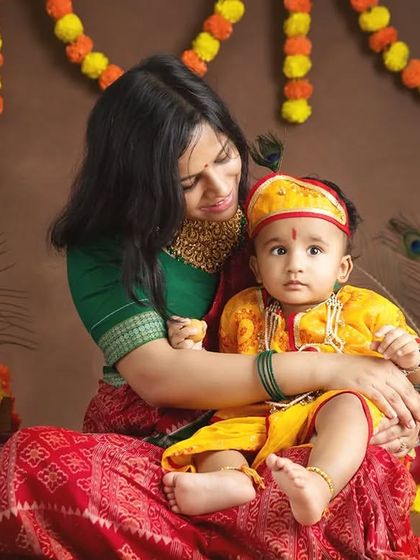 A mother dressed in traditional attire holds her baby, who is dressed as little Krishna for a Janmashtami themed shoot.