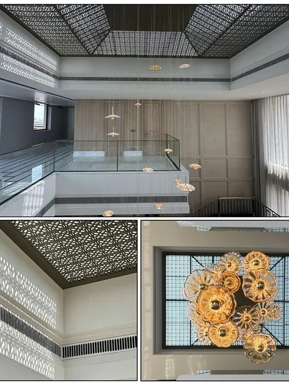In this triple-height space, a decorative jaali in the skylight creates captivating shadow patterns on the walls below. This detail, combined with a cascading chandelier, adds an artistic touch and enhances the grand ambiance of the home.