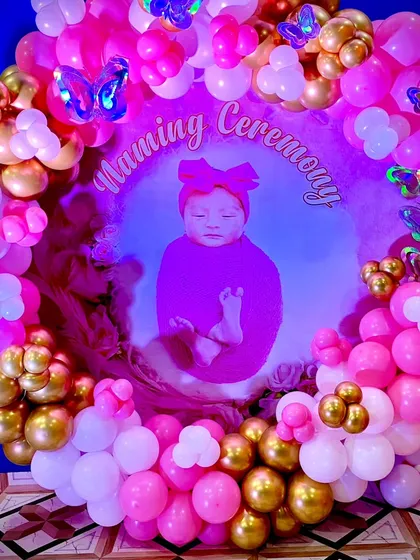 A close-up of the personalized Naming Ceremony backdrop. The baby's photo is surrounded by a dense ring of pink, white, and gold balloons with shimmering butterfly details.