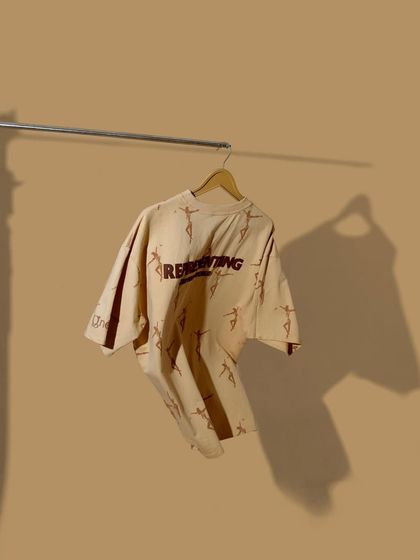 A minimalist, on-hanger shot of a beige t-shirt against a matching background. The use of a single, hard light source creates a distinct shadow, adding an artistic touch.