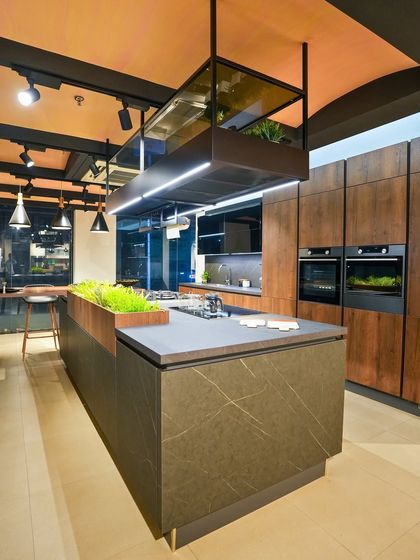 A luxurious kitchen display in Kolkata with dark wood cabinets, a veined stone-look island, and a suspended chimney with integrated lighting and plant holders.