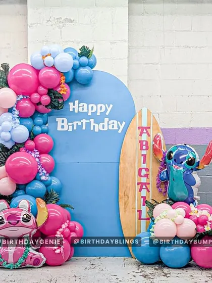 A cute Lilo & Stitch themed party with a tropical vibe. This setup includes cutouts of Stitch and Angel, a personalized surfboard, and a pink and blue balloon garland.