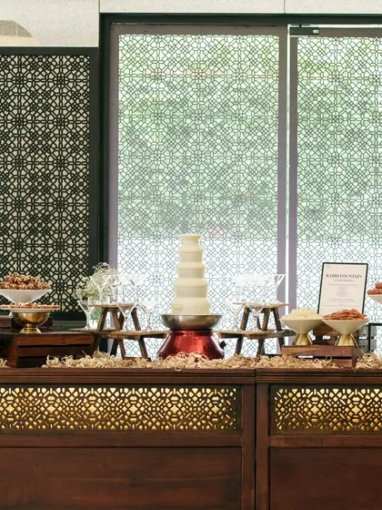 An elegant dessert buffet station with a white chocolate fountain as the centrepiece. The intricate woodwork of the counter and the symmetrical arrangement create a look of refined luxury.