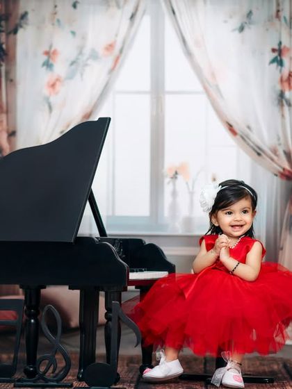 A joyful little girl in a red dress claps her hands with delight at her mini piano in our elegant, floral-themed studio.