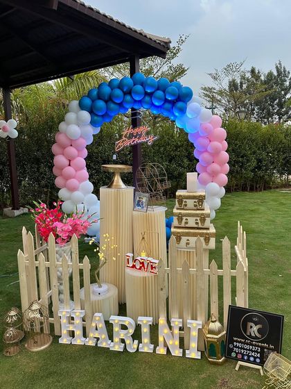 A lovely outdoor setup on a green lawn. The circular balloon arch, picket fence, and themed plinths create a beautiful focal point for a garden party.