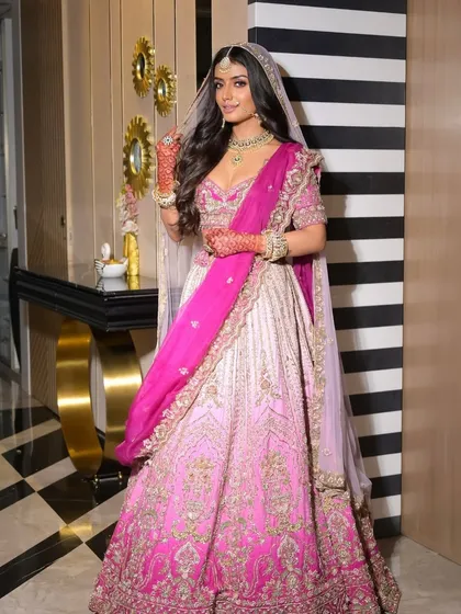 A full-length view of a new-age bridal look in a vibrant pink ombre lehenga. This image shows how the complete look comes together, with the makeup, hair, and outfit creating a cohesive and stunning vision. The makeup is kept fresh and radiant to balance the intricate outfit.