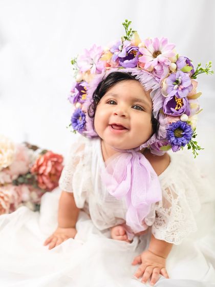 A beautiful eight-month-old baby smiles brightly, wearing an incredible floral bonnet. Milestone sessions are a chance to get creative with styling and props.