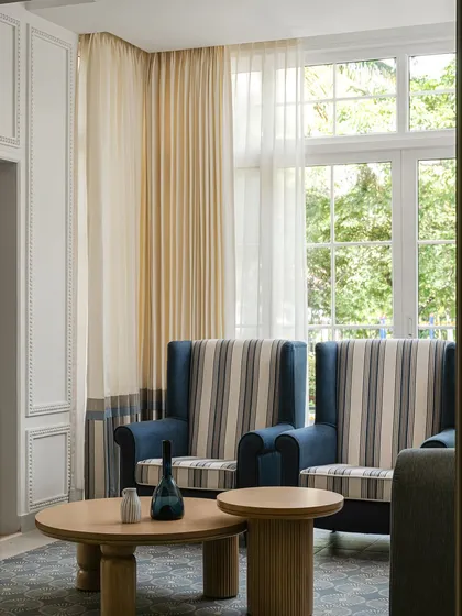 Sunlight streams into this serene sunroom, highlighting the classic striped wingback chairs and the gentle curves of the nested oak coffee tables. The design is a harmonious blend of classic and contemporary styles.
