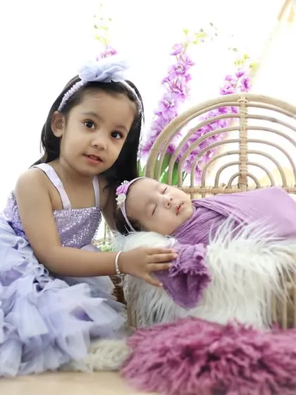 A big sister watches over her sleeping newborn sibling. The coordinated lavender outfits and floral background create a soft and dreamy sister-themed newborn shoot.