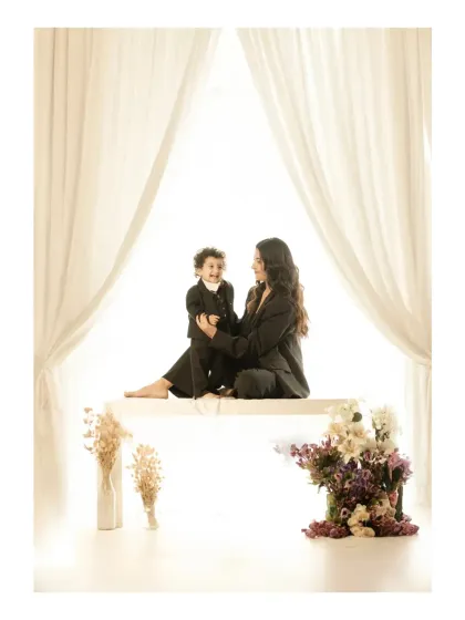 In a beautifully symmetric composition, a mother and son sit on a white bench, framed by flowing curtains. This indoor family shoot highlights their joyful interaction against a clean, minimalist backdrop.