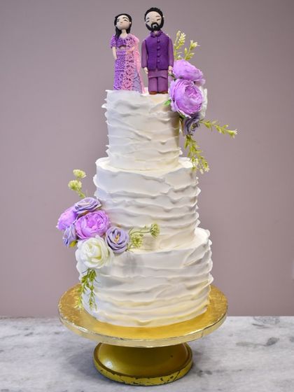 This four-tier wedding cake features a rustic, textured buttercream finish and is adorned with beautiful lavender-hued flowers. To make it truly personal, I sculpted custom edible figurines of the couple to sit atop their cake.