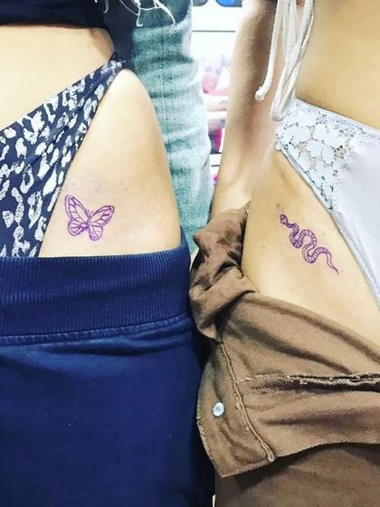 Friends showing their matching hip tattoos, one a butterfly and one a snake.