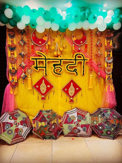 A colorful and traditional Mehendi backdrop. I used a bright yellow curtain, marigold flower lettering for "Mehndi," and decorative Rajasthani umbrellas to create a lively and festive photo corner.