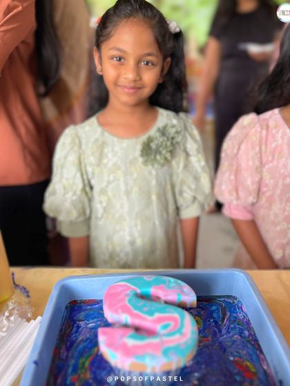 A young girl proudly stands behind her finished fluid art letter 'S', showcasing the beautiful pink and turquoise marbled effect she created.