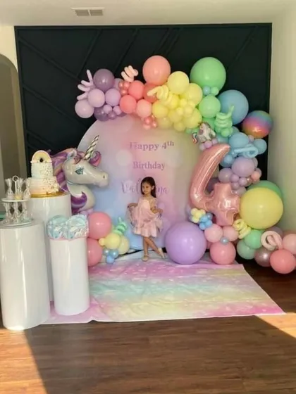 A dreamy unicorn and rainbow-themed 4th birthday setup, featuring pastel balloons, a unicorn figure, and pedestals for the cake display.