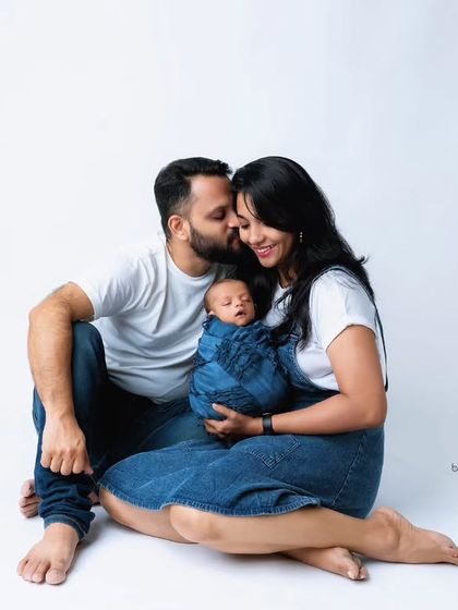 A happy family moment captured on a clean white background. The father kisses the mother's cheek as she holds their swaddled newborn, creating a relaxed and joyful portrait.