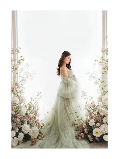 A full-length portrait showcasing a dreamy, ethereal look. The soft green gown and floral frame create a picture of a mother-to-be in her own secret garden.