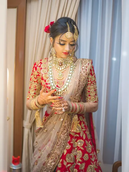A full-length portrait of a bride getting ready. This shot captures the final touches and the complete elegance of her red and gold wedding lehenga.