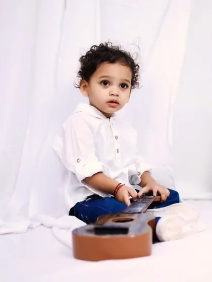 A toddler boy in a simple white shirt and jeans, playing with a toy guitar against a clean white background.