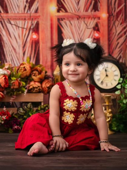 A rustic first birthday portrait in a warm, woody setting. The rich red dress provides a beautiful pop of color against the natural backdrop.