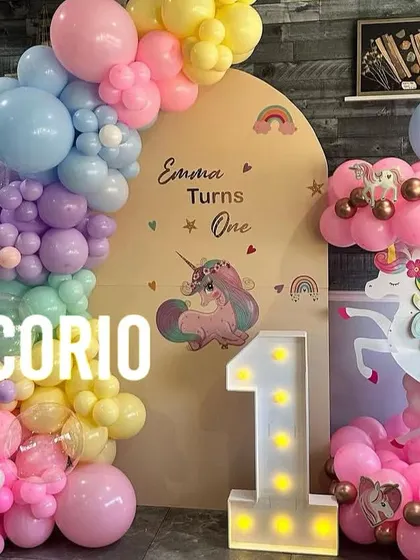 A magical unicorn-themed first birthday for 'Emma'. The setup includes a pastel rainbow balloon arch, a light-up number '1', and a unicorn-themed balloon column.