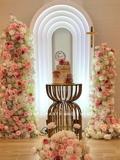 An elegant christening decoration for a girl. The centerpiece is a modern, lighted rainbow arch flanked by tall towers of pink and white flowers.