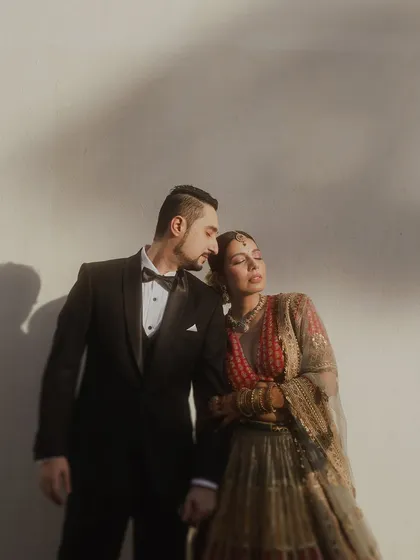 A moody, sun-drenched portrait where the groom in his tuxedo holds his bride in her stunning Sabyasachi lehenga. The play of light and shadow adds a dramatic, high-fashion feel to this wedding portrait.