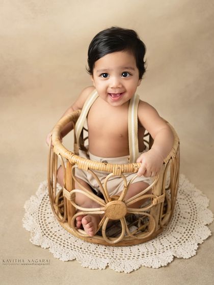 A ten-month-old boy sits happily in a wicker basket. His bright smile and playful pose are what make sitter sessions so much fun.