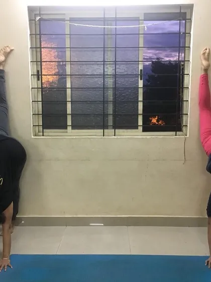 Two students practicing a wall-supported leg split, a great way to build flexibility for Hanumanasana.