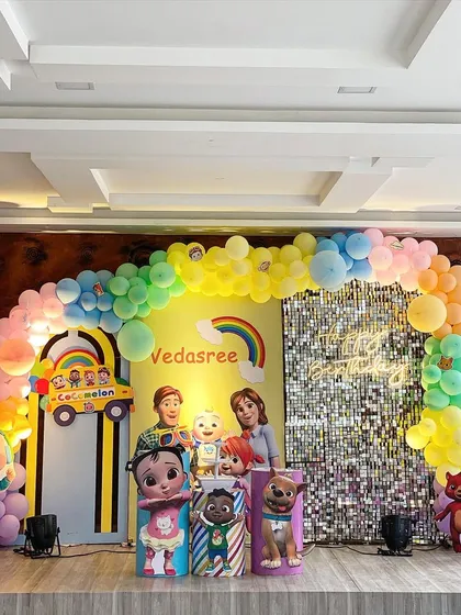 For the little ones, I design colorful themes like this Cocomelon party. It features a rainbow balloon arch, a silver shimmer wall, life-sized character cutouts of JJ and his friends, and a light-up marquee number '1' for a first birthday.