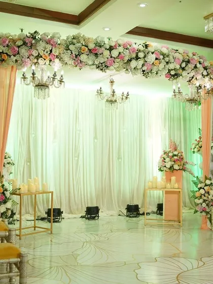 A lovely pastel-themed stage for a pre-wedding ceremony, featuring a floral arch, elegant drapes, and classic chandeliers for a soft, romantic look.