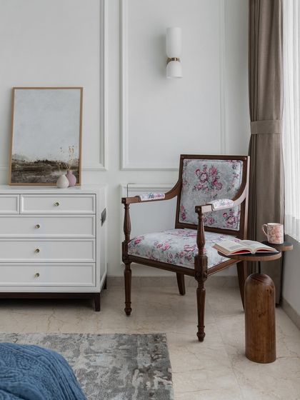 A quiet corner designed for peaceful moments. A vintage-style floral armchair is paired with a simple side table and a clean, white chest of drawers, creating a serene spot within a larger room.