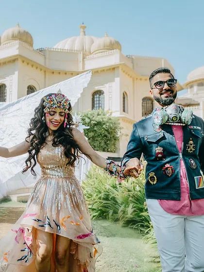 The happy couple, in their bold festival attire, walk hand-in-hand through the gardens of Udaivilas, with a winged performer on stilts in the background.