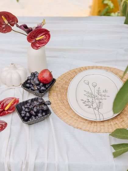 A close-up of the place setting on the Orchard Table. A beautifully illustrated plate sits on a woven placemat, surrounded by fresh grapes and anthurium petals.