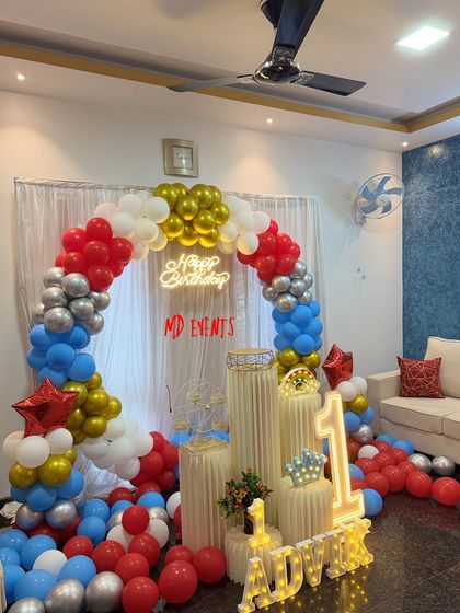 A classic first birthday balloon decoration for a home party. This setup features a circular arch of red, blue, silver, and gold balloons, a 'Happy Birthday' neon sign, and a light-up '1' and name display.