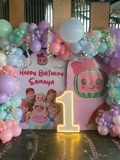 A Cocomelon first birthday party setup with a pastel color palette. The decor includes a custom backdrop with characters, a light-up number '1', and a full balloon arch in soft shades of purple, pink, and mint green.