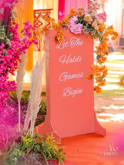 A closer view of the fun "Let the Haldi Games Begin" sign, adorned with bougainvillea and pampas grass.