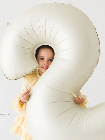 A playful portrait for a second birthday. The little girl peeks out from behind a giant number '2' balloon with a mischievous smile.