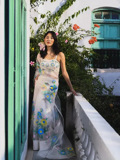 A full-length shot on a balcony, with the sheer, hand-painted saree catching the light beautifully. This image feels fresh, airy, and effortlessly elegant.