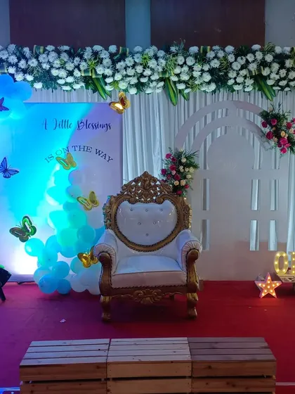 This baby shower stage combines modern and traditional touches. It has an elegant throne chair, a white floral garland overhead, and a backdrop with blue lighting and butterfly accents to welcome the new baby.
