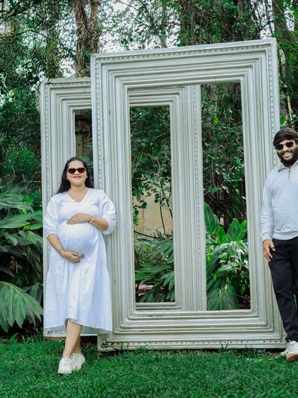 A fun and creative outdoor portrait of an expecting couple. They pose with large empty picture frames in a garden, creating a unique and playful composition.