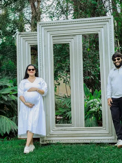 A fun and creative outdoor portrait of an expecting couple. They pose with large empty picture frames in a garden, creating a unique and playful composition.