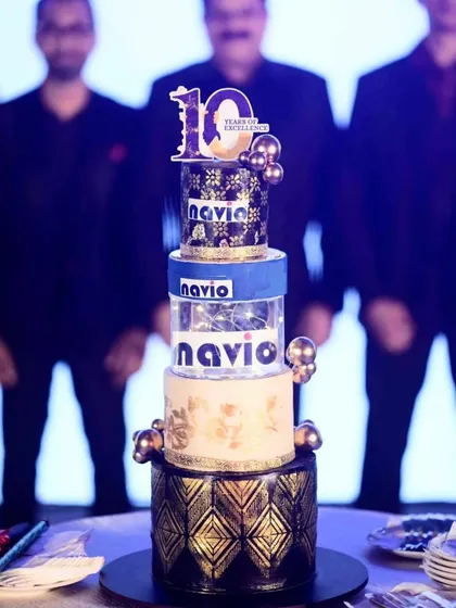 A towering four-tier cake for the 10th anniversary of the shipping company, NAVIO. This corporate cake features the company's branding, a black and gold color scheme, and an illuminated middle tier.