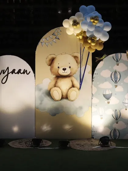 A lovely teddy bear themed backdrop for Avyaan's first birthday. The setup features three arched panels with a 3D teddy bear, hot air balloon prints, and personalized lettering.