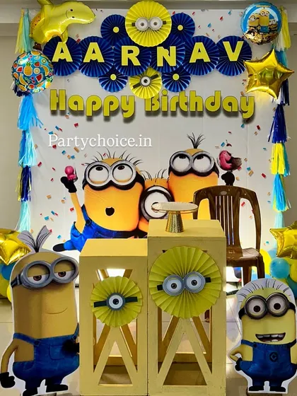 A fun-filled Minions party decoration. This setup is designed with minimal balloons, focusing on a large character backdrop, themed pedestals, paper fan decorations, and Minion cutouts.