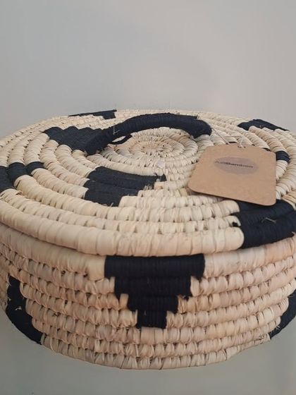 A round, lidded basket woven from palm leaf with a black geometric pattern. This decorative box is perfect for storing jewelry, trinkets, or other small treasures while adding a global-inspired accent to your decor.
